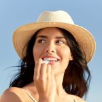 portret van gelukkig lachende jonge vrouw in bikini badpak en stro hoed lippenbalsem toe te passen op het strand