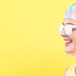 Vrouw in een kleurrijke pruik met koptelefoon luisteren naar muziek met bril op en shimmer powders 9 stacks van minerale make up bellapiere op haar gezicht kijkend naar links, geïsoleerd op een gele achtergrond