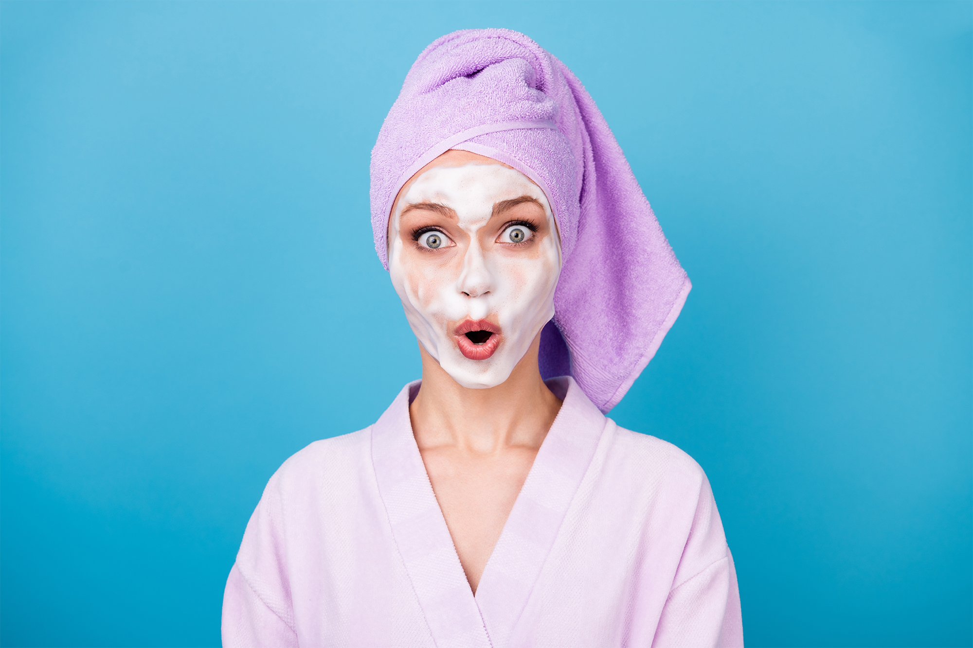 Foto van schattige vrouw met wit gezichtsmasker van 24nexx, draag paarse handdoek tulband badjas open mond, geïsoleerde blauwe kleur achtergrond