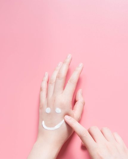 Mooie vrouw handen hydraterende crème toe te passen op roze achtergrond. Lachende emoji-vorm gemaakt van lotion. Verzorg je lichaamshuid. Gezondheidszorg en schoonheidsconcept. Bovenaanzicht. Detailopname.